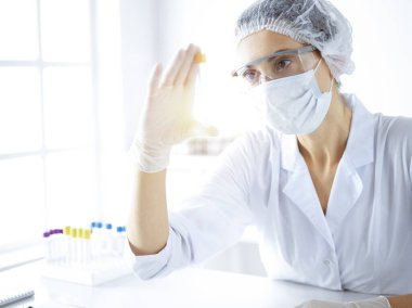 Koruyucu gözlüklü profesyonel kadın bilim adamı güneşli laboratuvarda reaktörlerle araştırma tüpü kullanıyor. Tıp ve bilim araştırmaları