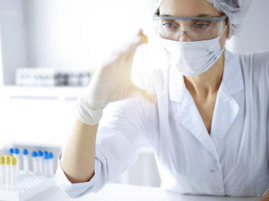 Koruyucu gözlüklü profesyonel kadın bilim adamı güneşli laboratuvarda reaktörlerle araştırma tüpü kullanıyor. Tıp ve bilim araştırmaları