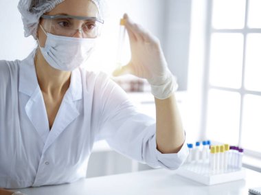 Koruyucu gözlüklü profesyonel kadın bilim adamı güneşli laboratuvarda reaktörlerle araştırma tüpü kullanıyor. Tıp ve bilim araştırmaları