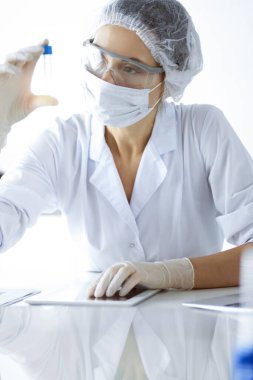 Koruyucu gözlüklü profesyonel kadın bilim adamı güneşli laboratuvarda reaktörlerle araştırma tüpü kullanıyor. Tıp ve bilim araştırmaları