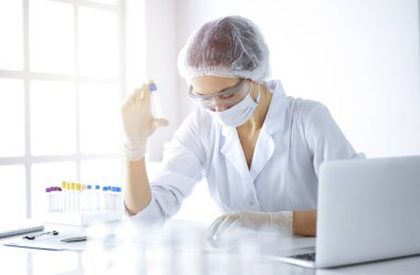 Koruyucu gözlüklü profesyonel kadın bilim adamı güneşli laboratuvarda reaktörlerle araştırma tüpü kullanıyor. Tıp ve bilim araştırmaları