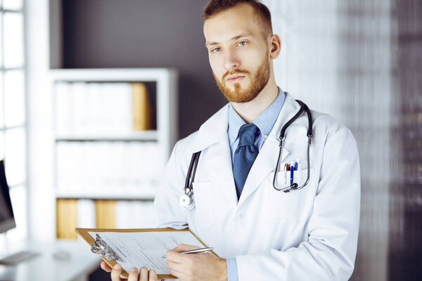 Friendly red-bearded doctor standing and writing with clipboard in clinic at his working place. Perfect medical service in hospital. Medicine concept