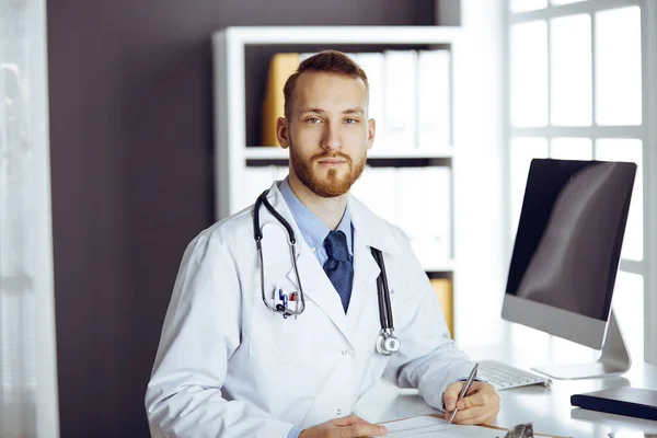 빨간 수염의 의사가 그의 직장 근처의 진료소에 바로 서 있습니다. 의사의 초상화. 의학과 건강 관리 개념 — 스톡 사진