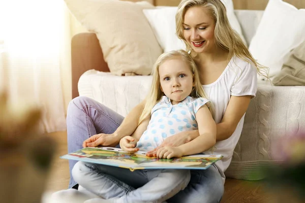 Happy family. Blonde young mother reading a book to her cute daughter while sitting at wooden floor in sunny room. Motherhood concept