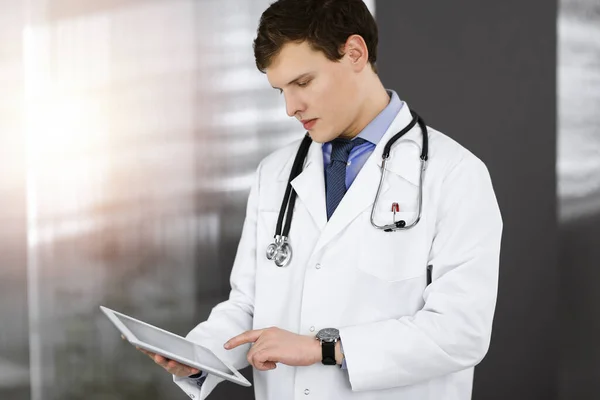 Mladý přátelský doktor kontroluje nějaké informace na svém počítači. Portrét profesionálního lékaře při práci na slunné klinice — Stock fotografie