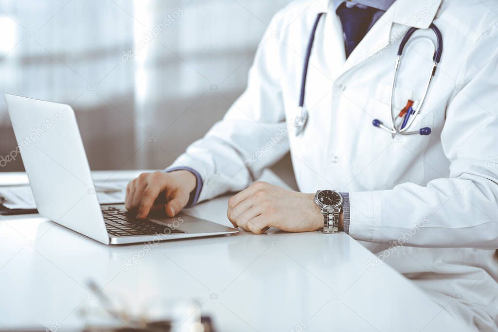 Unknown male doctor sitting and working with laptop in clinic at his working place, close-up. Young physician at work. Perfect medical service, medicine concept