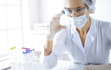 Koruyucu gözlüklü profesyonel kadın bilim adamı güneşli laboratuvarda reaktörlerle araştırma tüpü kullanıyor. Tıp ve bilim araştırmaları