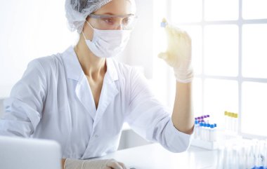 Koruyucu gözlüklü profesyonel kadın bilim adamı güneşli laboratuvarda reaktörlerle araştırma tüpü kullanıyor. Tıp ve bilim araştırmaları