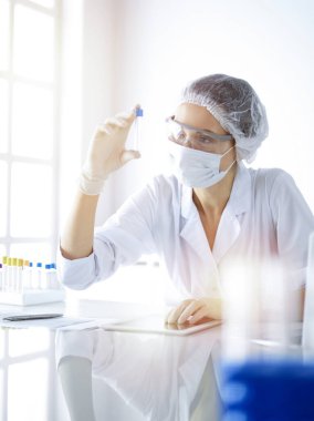 Koruyucu gözlüklü profesyonel kadın bilim adamı güneşli laboratuvarda reaktörlerle araştırma tüpü kullanıyor. Tıp ve bilim araştırmaları