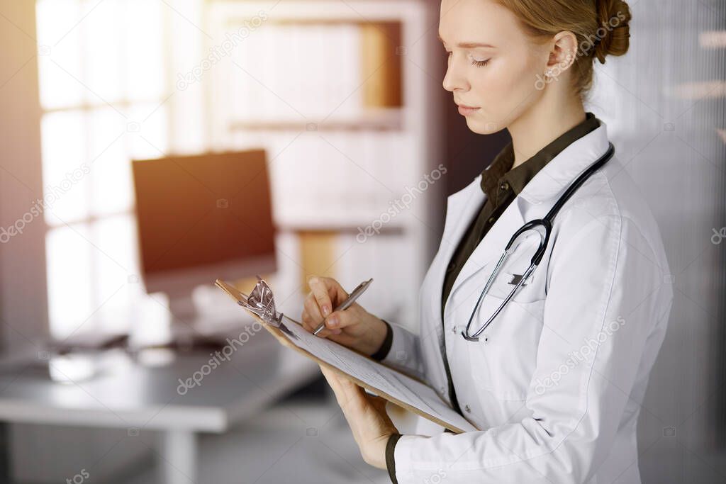 Cheerful smiling female doctor using clipboard in sunny clinic. Portrait of friendly physician woman at work. Perfect medical service in hospital. Medicine concept