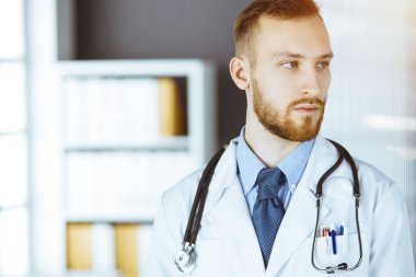 Kızıl sakallı doktor çalışma yerinin yanındaki güneşli klinikte dikiliyor. Hekim portresi. Tıp konsepti