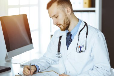 Dost canlısı kızıl sakallı doktor güneşli klinikteki panoda oturuyor ve yazıyor. Tıp konsepti