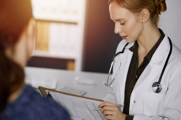 Alegre sorridente feminino médico e paciente mulher discutindo exame de saúde atual enquanto sentado na clínica ensolarada. Serviço médico perfeito no hospital. Conceito de medicina — Fotografia de Stock