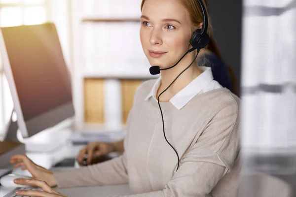 Femme d'affaires parlant par casque tout en étant assis dans un bureau ensoleillé. Centre d'appels et divers groupes de personnes en affaires — Photo