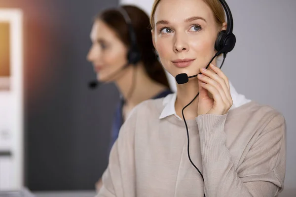 Femme d'affaires parlant par casque tout en étant assis dans un bureau ensoleillé. Centre d'appels et divers groupes de personnes en affaires — Photo