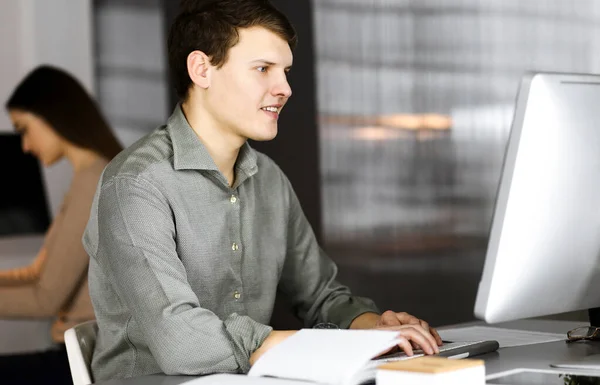 Темноволосый молодой бизнесмен и программист в зеленой рубашке усердно работает за компьютером, сидя за столом в современном кабинете с коллегой-женщиной на заднем плане. Концепция — стоковое фото
