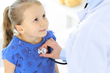 Doktor steteskopla küçük bir kızı muayene ediyor. Her zamanki tıbbi muayenede gülümseyen çocuk hasta. Tıp ve sağlık hizmetleri kavramları