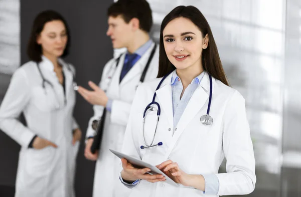 Usmívající se inteligentní lékařka drží v rukou tablet, zatímco ona stojí se svými kolegy na klinice. Lékaři v práci. Perfektní lékařská služba v nemocnici — Stock fotografie