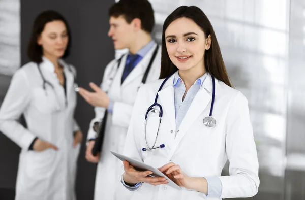 Gülümseyen zeki kadın-doktor, klinikte meslektaşlarıyla birlikte ayakta dururken elinde bir tablet bilgisayar tutuyor. Doktorlar iş başında. Hastanede mükemmel bir tıbbi hizmet.