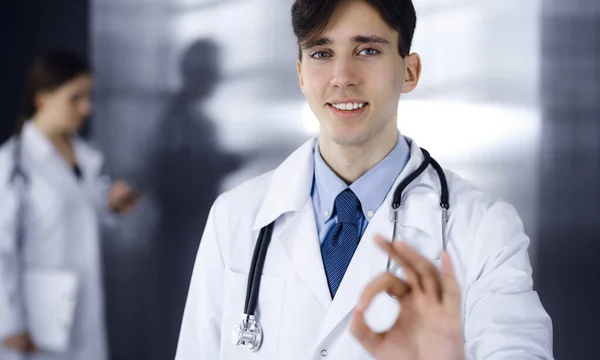 Freundlicher Mann-Arzt, der gerade steht und in der Klinik Ok-Zeichen zeigt. Die Kollegin hat es eilig im Hintergrund des Arztes. Medizinkonzept — Stockfoto