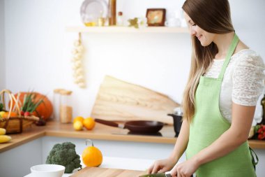 Güneşli mutfakta önlük giymiş güzel bir kadın yemek pişiriyor.