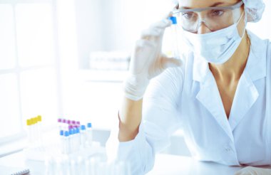 Koruyucu gözlüklü profesyonel kadın bilim adamı güneşli laboratuarda mavi tonda reaksiyonlarla araştırma tüpü kullanıyor. Tıp ve bilim araştırmaları