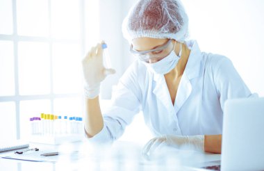 Koruyucu gözlüklü profesyonel kadın bilim adamı güneşli laboratuarda mavi tonda reaksiyonlarla araştırma tüpü kullanıyor. Tıp ve bilim araştırmaları