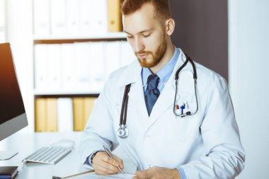 Dost canlısı kızıl sakallı doktor güneşli klinikteki panoda oturuyor ve yazıyor. Tıp konsepti