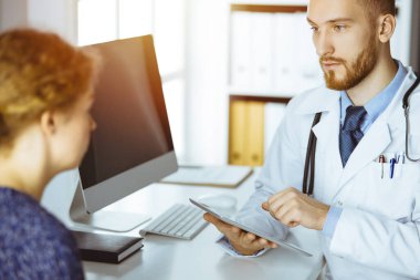 Dost canlısı kızıl sakallı doktor ve hasta kadın güneşli klinikte oturup tablet bilgisayar kullanırken sağlık muayenesini tartışıyorlar. Tıp konsepti