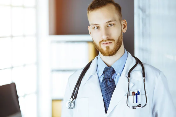Red-bearded doctor standing straight in sunny clinic near his working place. Portrait of physician. Medicine concept