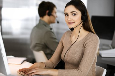 Modern bir ofiste meslektaşıyla birlikte masa başında otururken, kulaklıklı genç bir kız bir firma müşterisiyle konuşuyor. İş yerindeki çağrı merkezi operatörleri