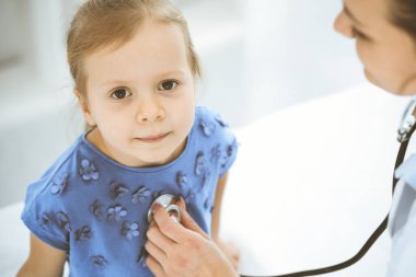 Doktor steteskopla küçük bir kızı muayene ediyor. Her zamanki tıbbi muayenede gülümseyen çocuk hasta. Tıp ve sağlık hizmetleri kavramları