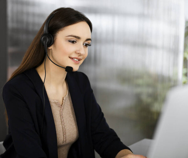 Молодая красивая женщина, одетая в наушники, слушает клиента компании, пока она сидит за столом, работая вместе с коллегой-мужчиной в современном офисе. Сосредоточься на женщине. Звонок