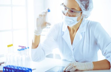 Koruyucu gözlüklü profesyonel kadın bilim adamı güneşli laboratuarda mavi tonda reaksiyonlarla araştırma tüpü kullanıyor. Tıp ve bilim araştırmaları