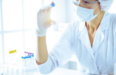 Koruyucu gözlüklü profesyonel kadın bilim adamı güneşli laboratuarda mavi tonda reaksiyonlarla araştırma tüpü kullanıyor. Tıp ve bilim araştırmaları