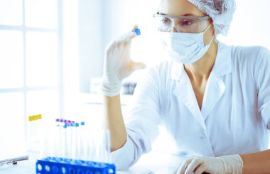 Koruyucu gözlüklü profesyonel kadın bilim adamı güneşli laboratuarda mavi tonda reaksiyonlarla araştırma tüpü kullanıyor. Tıp ve bilim araştırmaları