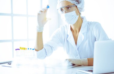 Koruyucu gözlüklü profesyonel kadın bilim adamı güneşli laboratuarda mavi tonda reaksiyonlarla araştırma tüpü kullanıyor. Tıp ve bilim araştırmaları