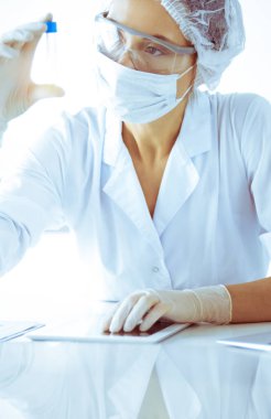 Koruyucu gözlüklü profesyonel kadın bilim adamı güneşli laboratuarda mavi tonda reaksiyonlarla araştırma tüpü kullanıyor. Tıp ve bilim araştırmaları