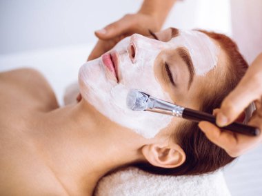 Güzel esmer kadın güneşli spa merkezinde kapalı gözlerle kozmetik maske takmaktan zevk alıyor. Rahatlatıcı tedavi ve ilaç konsepti