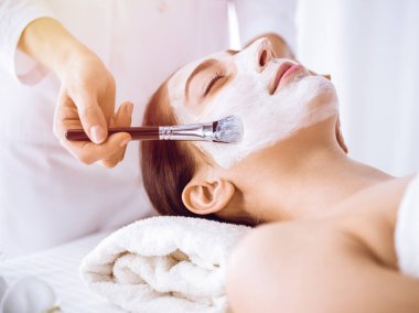 Güzel esmer kadın güneşli spa merkezinde kapalı gözlerle kozmetik maske takmaktan zevk alıyor. Rahatlatıcı tedavi ve ilaç konsepti