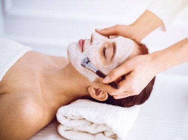 Güzel esmer kadın güneşli spa merkezinde kapalı gözlerle kozmetik maske takmaktan zevk alıyor. Rahatlatıcı tedavi ve ilaç konsepti