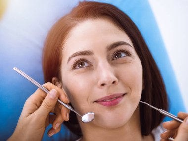 Güneşli diş kliniğinde dişçi tarafından muayene edilen gülümseyen esmer kadın. Hastanın ağzının yanında diş aletleri tutan bir doktorun elleri. Sağlıklı diş ve ilaç konsepti