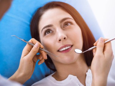 Güneşli diş kliniğinde dişçi tarafından muayene edilen gülümseyen esmer kadın. Hastanın ağzının yanında diş aletleri tutan bir doktorun elleri. Sağlıklı diş ve ilaç konsepti