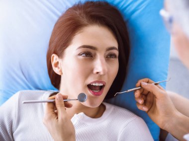 Güneşli diş kliniğinde dişçi tarafından muayene edilen gülümseyen esmer kadın. Hastanın ağzının yanında diş aletleri tutan bir doktorun elleri. Sağlıklı diş ve ilaç konsepti