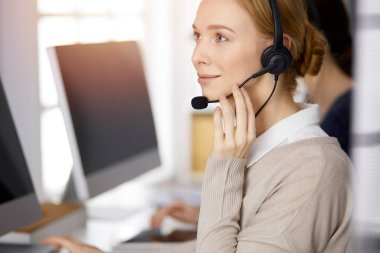 İş kadını güneşli bir ofiste otururken kulaklıkla konuşuyor. Çağrı merkezi ve çeşitli iş grupları