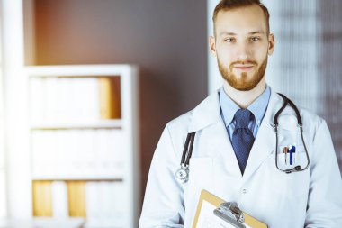 Dost canlısı kızıl sakallı doktor çalışma yerindeki güneşli klinikte panoya yazı yazıyor. Tıp konsepti