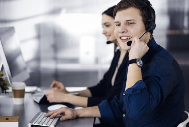 Modern bir ofiste masada otururken, kulaklıklı iki genç müşterilerle konuşuyor. Mavi gömlekli adama odaklan. İş yerindeki çağrı merkezi operatörleri