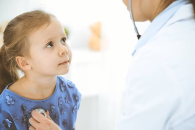 Doktor steteskopla küçük bir kızı muayene ediyor. Her zamanki tıbbi muayenede gülümseyen çocuk hasta. Tıp ve sağlık hizmetleri kavramları