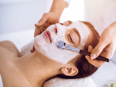 Güzel esmer kadın güneşli spa merkezinde kapalı gözlerle kozmetik maske takmaktan zevk alıyor. Rahatlatıcı tedavi ve ilaç konsepti