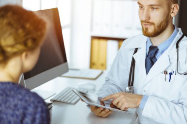 Dost canlısı kızıl sakallı doktor ve hasta kadın güneşli klinikte oturup tablet bilgisayar kullanırken sağlık muayenesini tartışıyorlar. Tıp konsepti